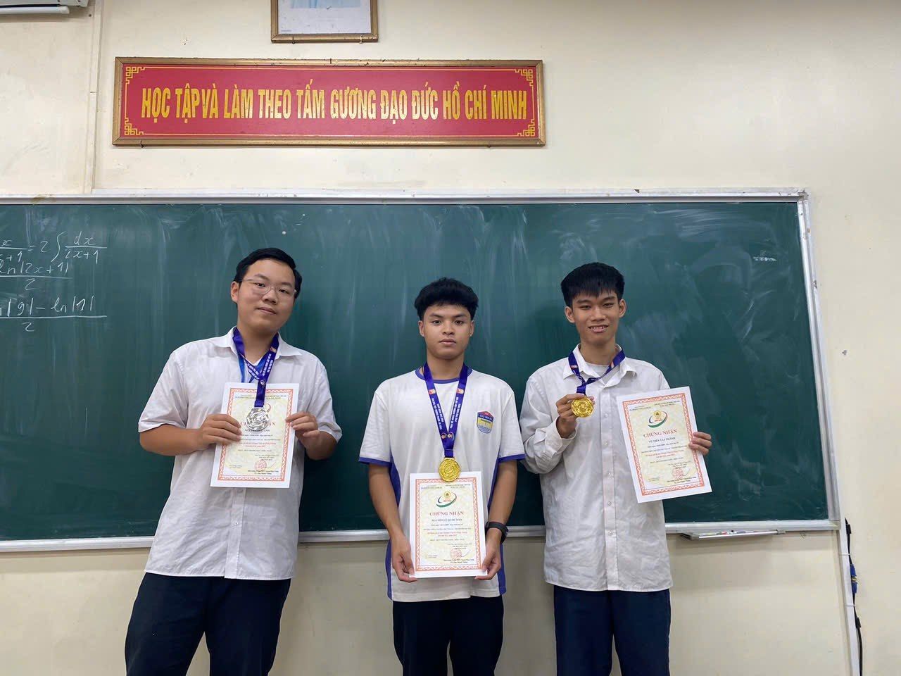 Three Chu Van An students holding certificates and medals in front of a blackboard.