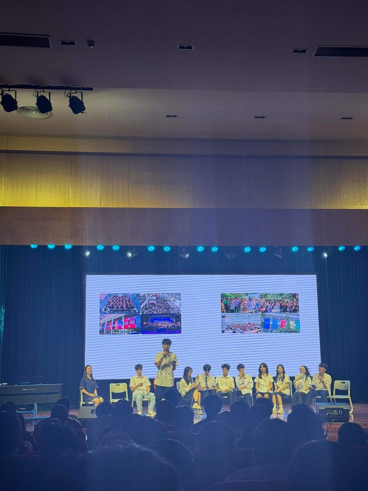 Students presenting on a stage with slides on a large screen.