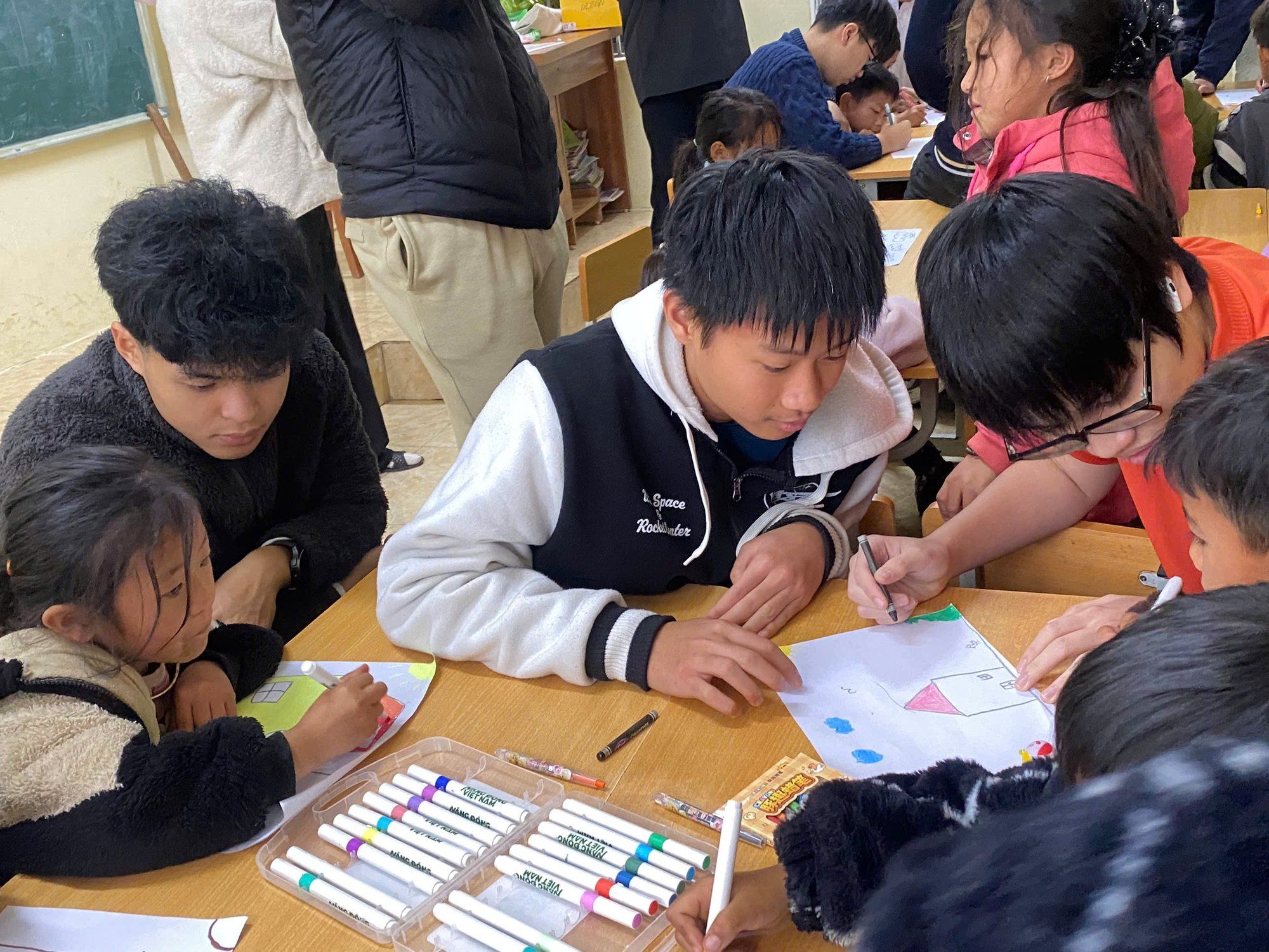 Volunteers and children drawing together at a classroom table.