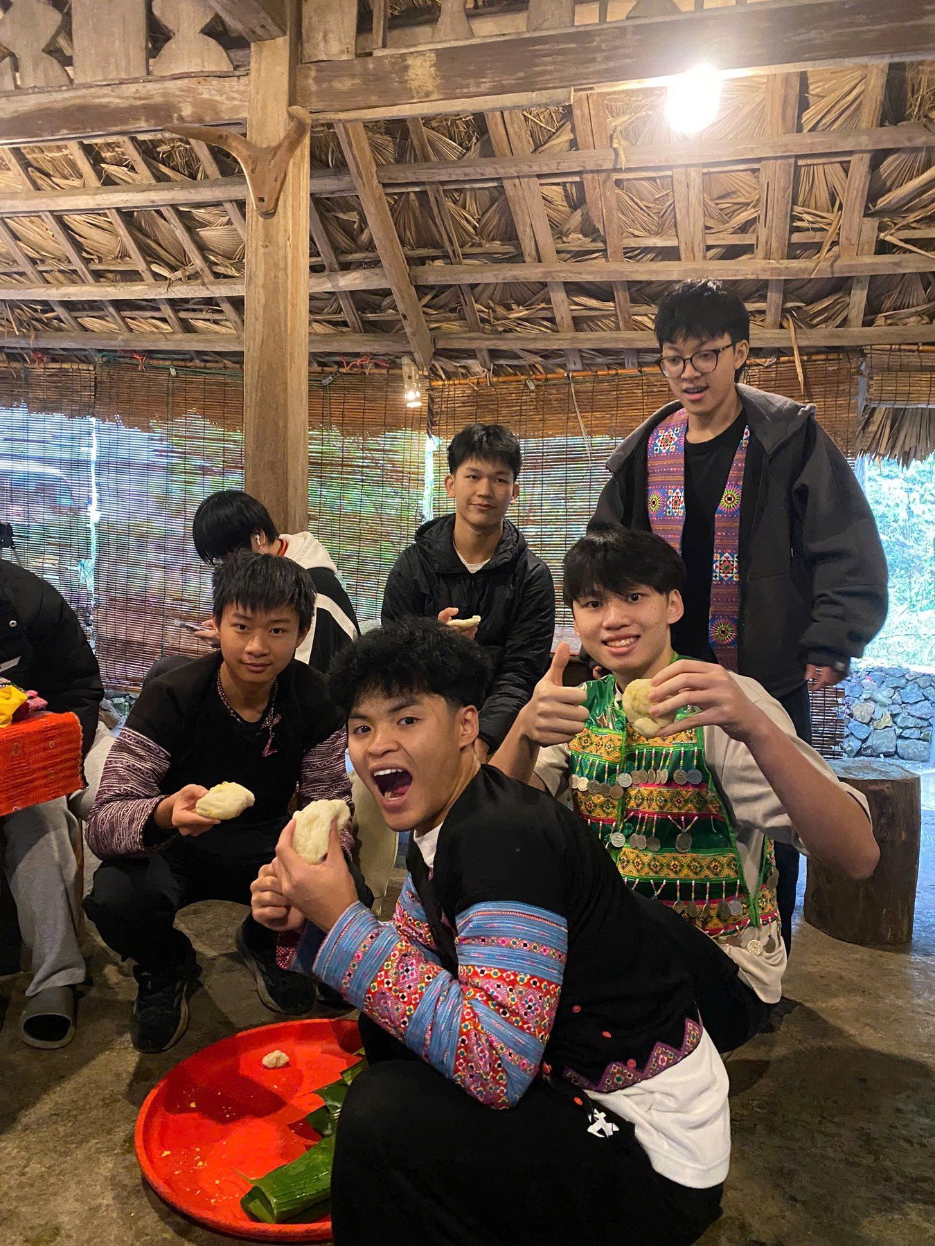 Bao and volunteers sharing food with local students under a wooden roof.